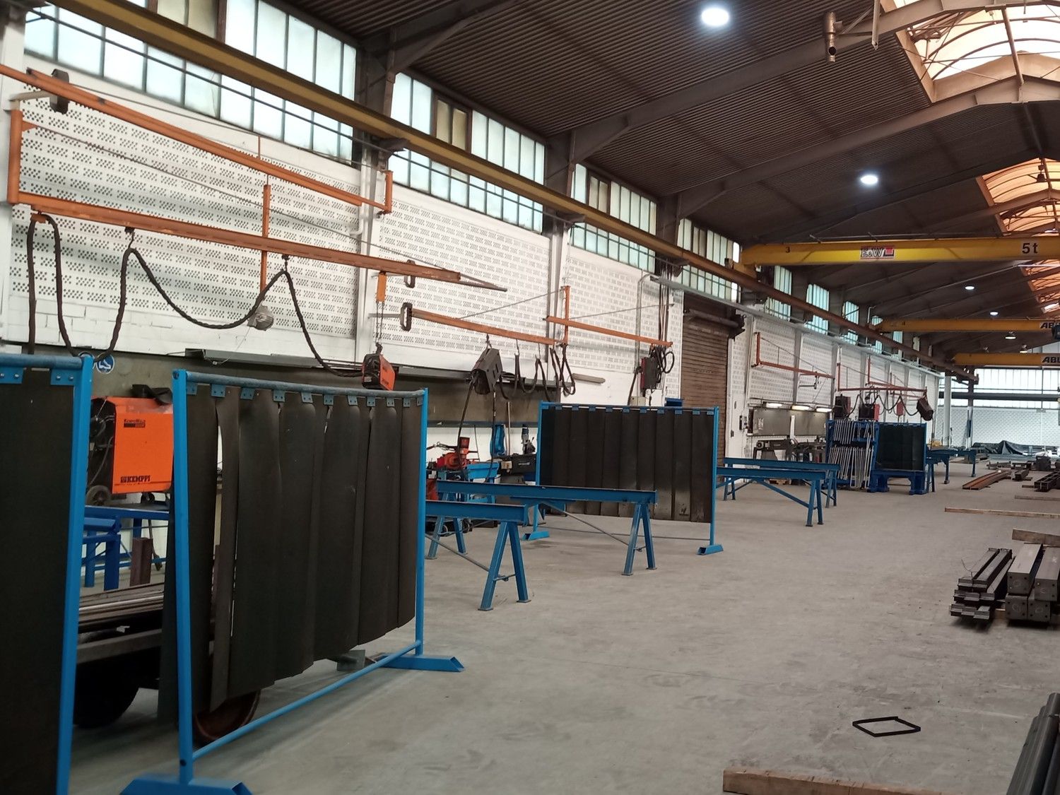 Industriehalle mit mehreren vormontierten Stahlrahmen, Metallbauteilen auf dem Boden und einem gelben Brückenkran im Hintergrund