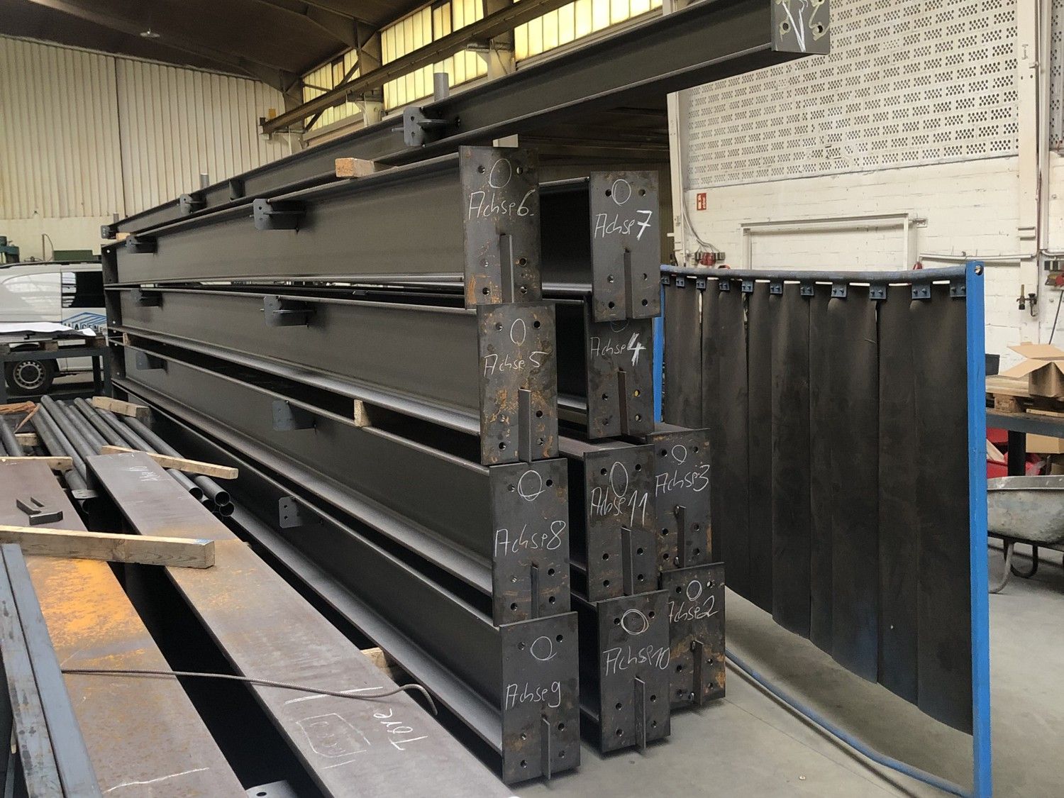 Bild zeigt eine große Industriehalle mit mehreren aufgestellten Stahlrahmen‑Konstruktionen, im Hintergrund ist ein Mobilkran erkennbar, im Vordergrund liegen Metallbauteile auf dem Boden.