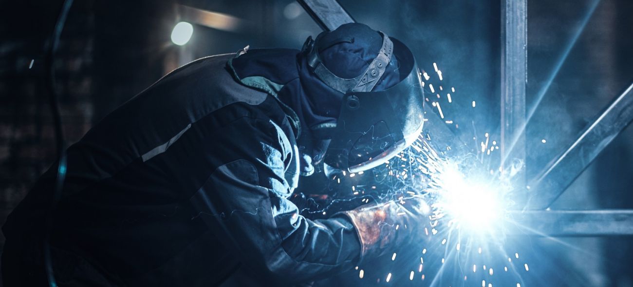 Ein Schweißer in Schutzkleidung und Helm arbeitet an einem Stahlgestell – Funken fliegen.